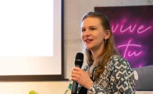 Frau mit Mikrofon spricht vor Publikum, im Hintergrund Leinwand und rosa Neonschrift „vive tu".