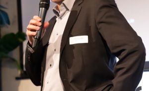 Mann mit Brille im Anzug hält Mikrofon, spricht vor Leinwand mit deutschem Text, trägt Namensschild.