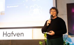 Person mit Mikrofon spricht vor Publikum, im Hintergrund Präsentationsfolie und Schild mit Aufschrift „Hafven".