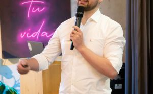 Mann in weißem Hemd und blauer Hose hält Mikrofon bei Vortrag. Im Hintergrund Neonschrift „vive tu vida".