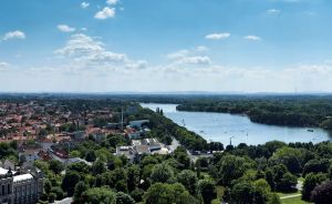 Luftaufnahme einer Stadt mit rotem Ziegeldächern, einem Fluss/See, umgeben von Grünflächen unter blauem Himmel.