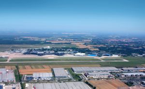 Luftaufnahme eines Flughafens mit Landebahn, Terminals und Hangars, umgeben von Feldern und Industriegebäuden.