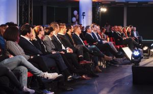 Publikum sitzt in Reihen bei einer Veranstaltung, beleuchtet von Scheinwerfern, im Vordergrund ein Bühnenstrahler.