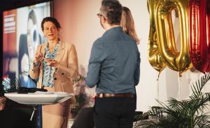 Frau spricht an Stehtisch, Mann steht ihr gegenüber, goldene Luftballons mit "40" im Hintergrund, Präsentationsscreen.