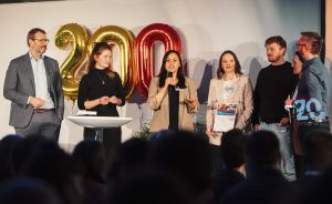 Sechs Personen auf einer Bühne, eine spricht ins Mikrofon, dahinter goldene Luftballons in Form von „200".