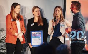 Drei Frauen auf einer Bühne, eine hält eine Urkunde, eine spricht ins Mikrofon, daneben ein Mann.