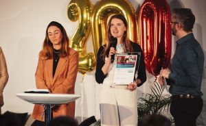 Frau spricht mit Mikrofon, hält Urkunde, neben ihr zwei Personen, im Hintergrund goldene Ballons „2000".