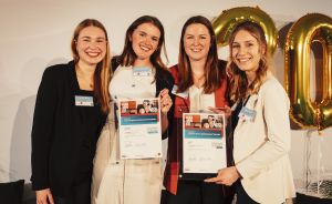 Vier junge Frauen stehen lächelnd nebeneinander, zwei halten Urkunden. Im Hintergrund goldene Luftballons.