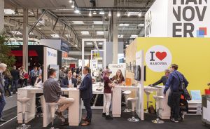 Messestand mit "I ❤ Hanover"-Schild und "HANNOVER"-Banner. Mehrere Personen stehen und sitzen an weißen Stehtischen.