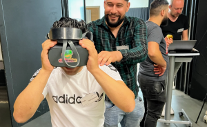 Mann mit Adidas-Shirt trägt VR-Brille, daneben stehender Mann in kariertem Hemd lächelt. Im Hintergrund zwei weitere Personen.