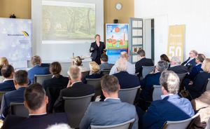 Eine Frau präsentiert vor Publikum in einem hellen Konferenzraum, im Hintergrund Leinwand und Roll-ups.