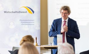 Mann im Anzug spricht vor Publikum bei WirtschaftsDienst-Veranstaltung, im Hintergrund Banner mit Hashtags.