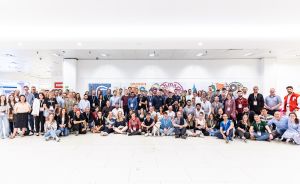 Große Gruppe von ca. 100 Personen posiert für ein Gruppenfoto in einem hellen Innenraum mit bunter Wandgestaltung.