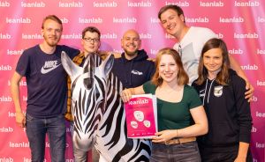 Sechs Personen posieren vor pinkfarbener Leanlab-Wand mit Zebrафигур und einem Leanlab-Plakat.