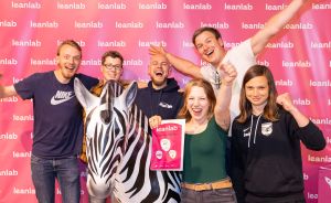Sechs Personen posieren lachend vor einer Leanlab-Wand mit einer Zebra-Figur. Eine Frau hält ein Leanlab-Schild.