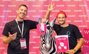 Zwei Männer posieren lächelnd vor pinker Leanlab-Wand mit Zebra-Statue und Urkunde „1. Platz".