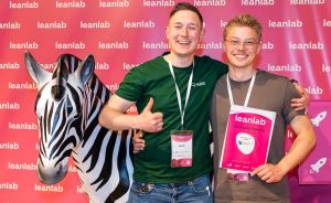 Zwei junge Männer lächeln vor leanlab-Hintergrund. Einer hält ein Zertifikat, neben ihnen ein Zebrakopf.
