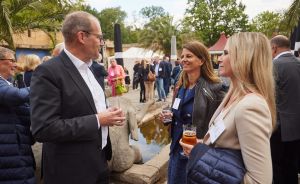 Drei Personen mit Getränken im Gespräch bei einer Outdoor-Veranstaltung, im Hintergrund weitere Gäste.