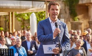 Mann im blauen Anzug spricht an einem Mikrofon vor einer Menschenmenge im Freien, hält ein Dokument in der Hand.