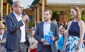 Drei Personen stehen auf einer Bühne. Ein Mann spricht ins Mikrofon, daneben ein Mann mit Mappe und eine Frau.
