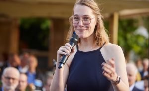 Junge Frau mit Brille und blondem Haar spricht in ein Mikrofon vor Publikum im Freien.