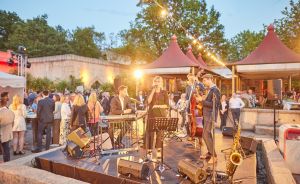 Eine Band spielt auf einer Bühne im Freien: Sängerin, Gitarrist, Kontrabassist, Saxofonist. Im Hintergrund feiernde Gäste.
