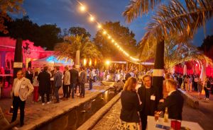 Abendliche Outdoor-Veranstaltung mit vielen Gästen, Lichterketten, Palmen und roter Beleuchtung im Hintergrund.