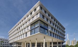 Modernes mehrstöckiges Bürogebäude mit Glasfassade und Betonsäulen unter blauem Himmel, Industrieschornsteine im Hintergrund.