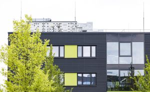 Modernes dunkles Bürogebäude mit gelb-grünen Fensterelementen, Lüftungsanlagen auf dem Dach, davor grüne Bäume.