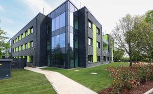 Modernes Bürogebäude mit schwarzer Metallfassade, grünen Akzenten und großen Glasfronten, umgeben von gepflegtem Rasen.