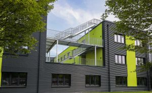 Modernes Gebäude mit schwarzer Fassade, grünen Akzenten und Balkonen, umgeben von Bäumen bei blauem Himmel.