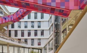 Modernes Atrium mit pixelgemustertem, rot-violettem Textilband über mehreren Stockwerken, Holztreppe im Vordergrund.