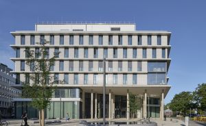 Modernes mehrstöckiges Bürogebäude mit Glasfassade, Säulen im Erdgeschoss, Bäumen und Platz davor, blauer Himmel.