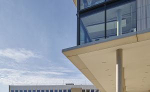 Modernes Gebäude mit Glasfassade und Überdachung, dahinter mehrstöckiges Bürogebäude, offener Platz mit Betonsteinen.