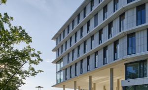 Modernes mehrstöckiges Bürogebäude mit Glasfassade, Arkaden im Erdgeschoss, Grünfläche und Fußgänger mit Fahrrad.