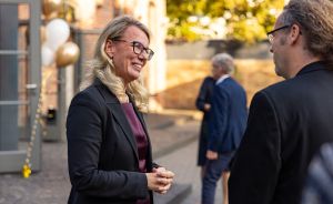 Frau mit Brille und schwarzem Blazer lacht im Gespräch mit Mann, im Hintergrund Person und gold-weiße Luftballons.