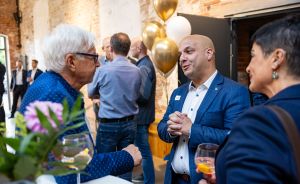 Drei Personen im Gespräch bei einer Veranstaltung mit gold-weißen Luftballons. Im Hintergrund weitere Gäste.