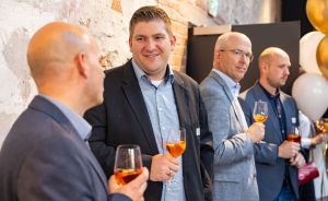Vier Männer in Businesskleidung stehen bei einer Veranstaltung und halten Weingläser mit orangefarbenem Getränk.
