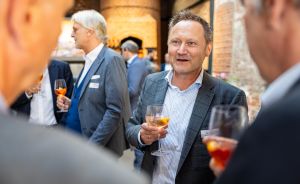 Mehrere Männer in Anzügen bei einer Networking-Veranstaltung, halten Cocktailgläser, im Hintergrund Backsteinwand.