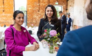 Zwei Frauen an einem Stehtisch mit Blumengesteck in einer Veranstaltungslocation aus rotem Backsteinmauerwerk.