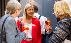 Drei Personen unterhalten sich bei einer Veranstaltung, zwei halten Aperol-Spritz-Gläser, eine Frau trägt rotes Jacket.