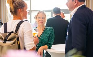 Drei Personen unterhalten sich bei einer Veranstaltung. Frau in grünem Kleid hält ein Glas. Im Hintergrund weitere Gäste.
