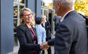 Zwei Personen in Businesskleidung geben sich vor einem Gebäude die Hand. Im Hintergrund stehen weitere Personen.