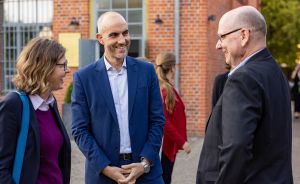 Drei Personen in Businesskleidung unterhalten sich lachend vor einem Backsteingebäude im Freien.