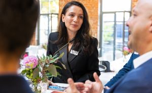 Frau in schwarzem Blazer mit Namensschild spricht mit zwei Personen an einem Tisch mit Blumen.