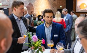 Zwei Männer in Jackets stehen an einem Stehtisch mit Blumen und halten Cocktailgläser. Im Hintergrund weitere Gäste.