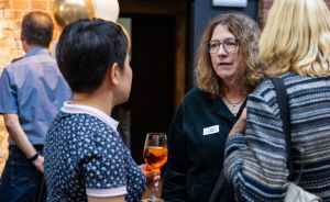 Drei Frauen im Gespräch bei einer Veranstaltung. Eine hält ein Glas Wein. Im Hintergrund ein Mann und Luftballons.