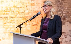 Blonde Frau mit Brille spricht lächelnd an einem Rednerpult mit Mikrofon vor einer Backsteinwand.
