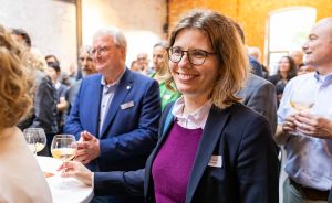 Frau mit Brille, Blazer und lila Pullover hält Weinglas bei einer Veranstaltung; mehrere Personen im Hintergrund.