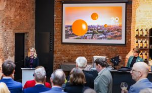 Frau spricht an Rednerpult vor Publikum, Leinwand zeigt „29 Years of Impulses" mit orangenen Kugeln über Stadtpanorama.